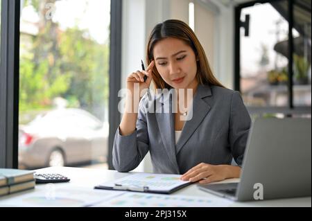 Focused and thoughtful millennial Asian businesswoman analyzing financial data on reports at her desk. Stock Photo