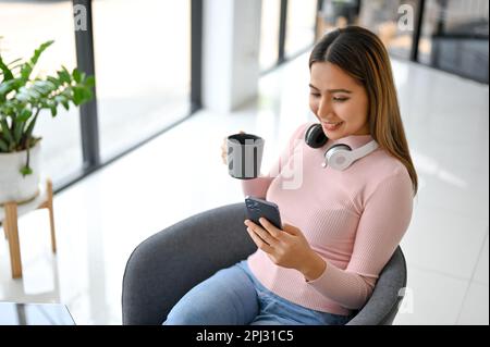 Charming and happy millennial Asian woman enjoying with her coffee and using her phone while relaxing in the modern coffee shop. Stock Photo