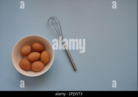 Eggs in white bowl and whisk on blue background - top view Stock Photo