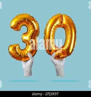 two hands in black and white holding two golden number-shaped balloons forming the number 30 on a pale blue background Stock Photo