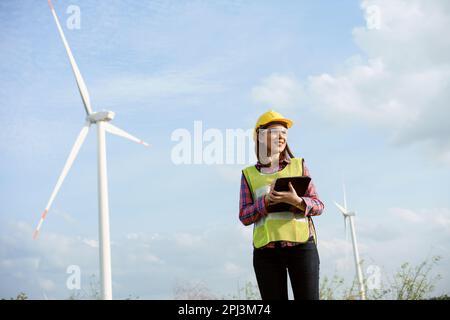 Asian woman in yellow helmet working with digital tablet at renewable energy farm. Female inspector controlling functioning of wind turbines outdoors. Stock Photo