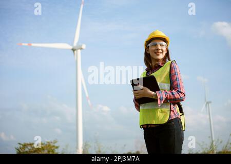 Asian woman in yellow helmet working with digital tablet at renewable energy farm. Female inspector controlling functioning of wind turbines outdoors. Stock Photo