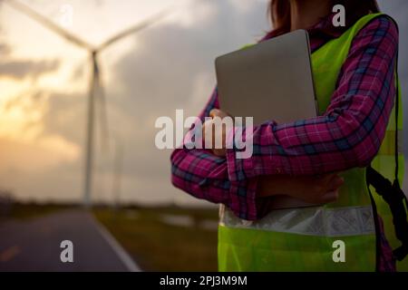 Asian woman working with laptop at renewable energy farm. Female inspector controlling functioning of wind turbines outdoors. Stock Photo