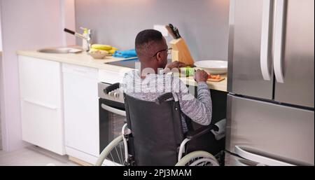 Young Disabled Man Sitting On Wheel Chair Preparing Food In Kitchen Stock Photo