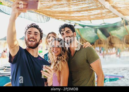 Four young friends, two men and two women, hugging and taking a selfie under a canopy on the beach, wearing summer outfits. Stock Photo
