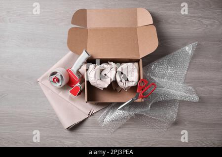 Open box with wrapped items, adhesive tape, scissors, paper and bubble wrap on wooden table, flat lay Stock Photo