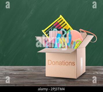 Donation box with different school stationery on wooden table near chalkboard Stock Photo