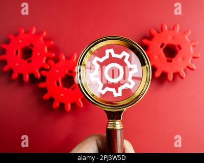 Digital gear wheel symbol appear on magnifying glass lens in businessman's hand while looking through on red gears on red background. Business strateg Stock Photo