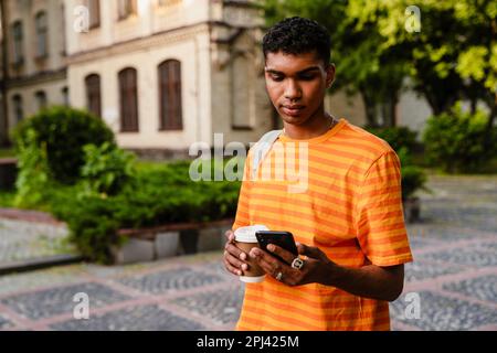 Young african man student in casual wear using smartphone and drinking coffee while standing outdoors in university campus Stock Photo