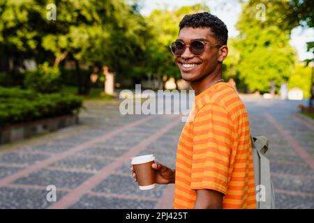 Young smiling african man student with backpack drinking coffee while standing outdoors Stock Photo