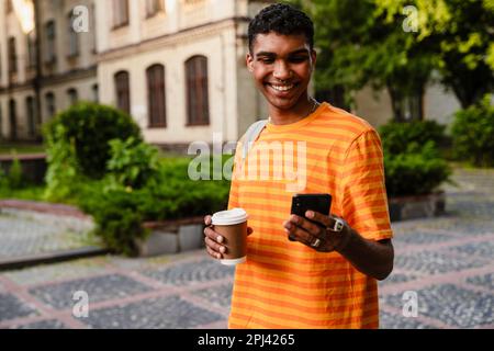 Smiling young african man student in casual wear using smartphone and drinking coffee while standing outdoors in university campus Stock Photo