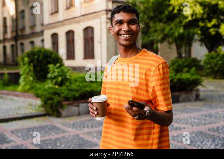 Smiling young african man student in casual wear using smartphone and drinking coffee while standing outdoors in university campus Stock Photo