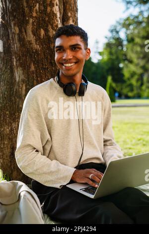 Young black man smiling and using laptop while sitting under tree in park Stock Photo