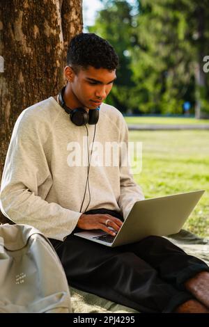 Young black man smiling and using laptop while sitting under tree in park Stock Photo