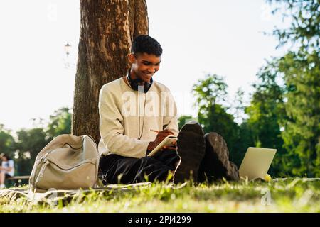 Young black man studying and taking notes while sitting under tree in park Stock Photo
