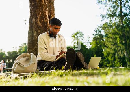 Young black man studying and taking notes while sitting under tree in park Stock Photo