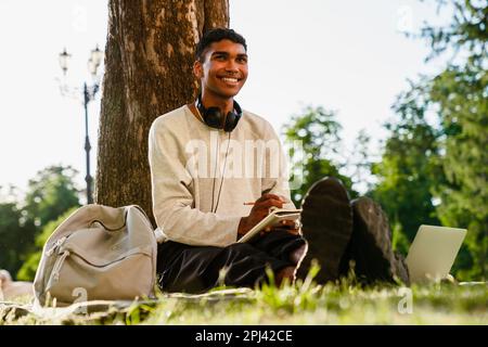 Young black man studying and taking notes while sitting under tree in park Stock Photo