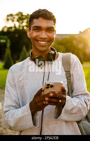 Young black man with headphones and backpack using mobile phone while walking by college outdoors Stock Photo
