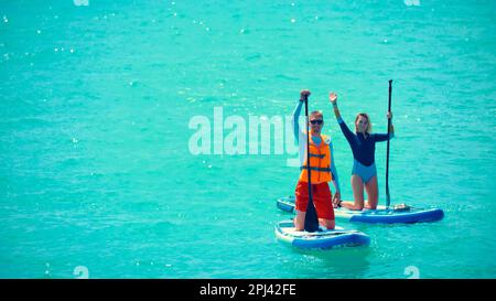 Man and woman rowing on stand up paddle boards. Young man dressed in swimsuit and lifevest and young woman waving on stand up paddle board. Summer out Stock Photo