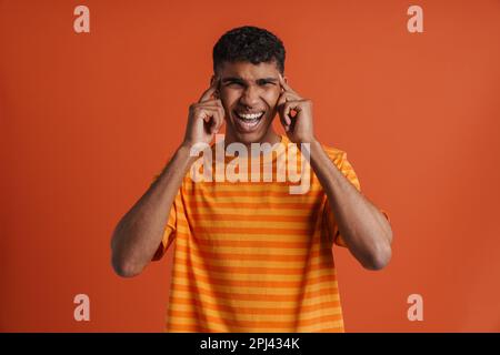 Young handsome tense squinting african man with opened mouth, touching his temples and looking at camera , while standing over isolated orange backgro Stock Photo