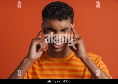 Young handsome tense african man with closed eyes and clenched teeth touching his temples, while standing over isolated orange background Stock Photo