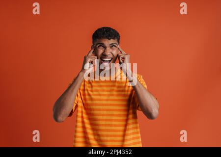 Young handsome tense squinting african man with opened mouth, touching his temples and looking upwards , while standing over isolated orange backgroun Stock Photo