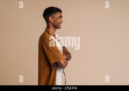 Profile portrait of young handsome smiling african man with piercing with folded arms, standing with his side over isolated beige background Stock Photo