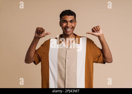 Young handsome smiling happy african man with piercing pointing on himself with both thumbs and looking at camera, while standing over isolated beige Stock Photo
