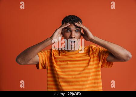 Young handsome shocked african man with piercing with opened mouth holding his head and looking at camera, while standing over isolated orange backgro Stock Photo