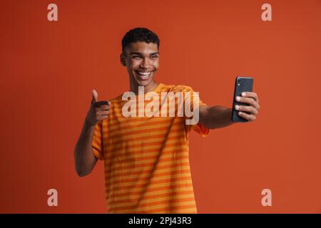 Young handsome happy smiling african man with piercing doing gun gesture and taking selfie on phone, while standing over isolated orange background Stock Photo