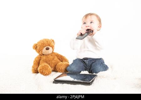 Stock studio photo with the white background of a baby's face with a mobile in his mouth next to a teddy bear and a tablet. Stock Photo