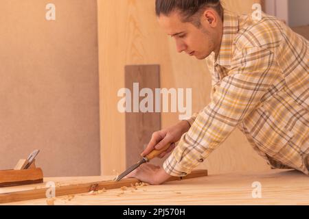Side view of attentive male woodworker carving wood with rasp near jack plane on workbench while working in modern workshop against boards of lumber Stock Photo