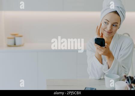 Thoughtful beautiful young woman applies face cream touches cheeks gently holds mirror looks aside dressed in soft bathrobe poses against kitchen inte Stock Photo