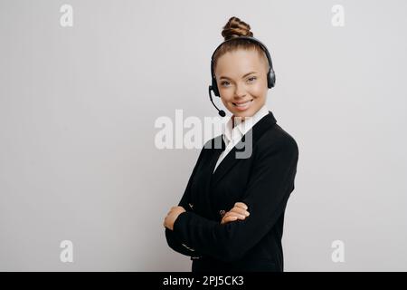 Female technical support worker in dark suit with black wireless headset standing sideways with her arms crossed and helping caller to resolve issues, Stock Photo