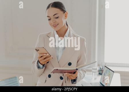 Successful business lady looking at smartphone screen with notepad in hand, reading last news or checking email. Smiling female employee in beige form Stock Photo