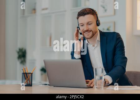 Happy successful businessman using laptop and typing on keyboard while sitting at the table and doing video call with headset, taking part in web conf Stock Photo