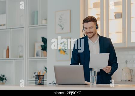 Happy young businessman wearing suit and headset working remotely from home while using video communication with colleagues on laptop, holding documen Stock Photo