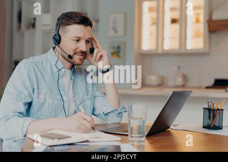 Happy bearded man with headset with microphone looking at laptop screen and making notes during distance education, sitting at his apartment while wor Stock Photo
