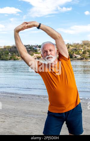 Happy energetic senior retired man stretching on the beach, doing side bend exercises by the river. Stock Photo