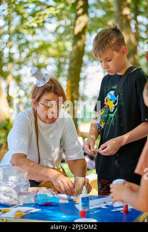 Svetlogorsk, Russia - 13.08.2022 - Funny adult woman in small paper hat teaches young guy make paper craft paper and scrapbooking at outdoor art festi Stock Photo