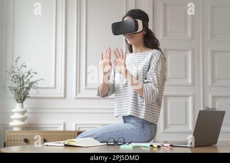 Young excited woman freelancer sitting on top of desk wearing VR glasses, hands raised in air seemingly against invisible wall in 3D game while intera Stock Photo