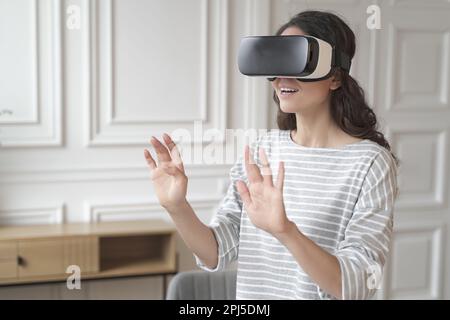 Young impressed brunette businesswoman interacting with virtual reality wearing VR headset glasses while relaxing between work online from home. Exite Stock Photo
