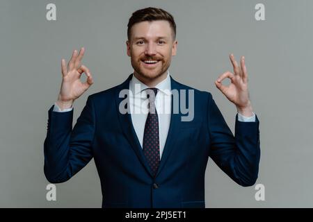 Successful bearded businessman shows ok sign with both hands dressed in formal clothes smiles pleasantly expresses approval recommends something isola Stock Photo