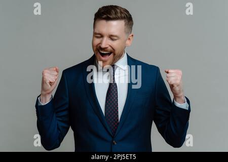 Cheerful excited bearded businessman makes yes gesture clenches fists feels like triumphant wears formal suit with tie celebrates success isolated ove Stock Photo