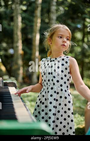 Svetlogorsk, Russia - 13.08.2022 - Cute little girl in polka dot dress playing piano outdoor in public park, outdoor art hobby of young girl, green fo Stock Photo