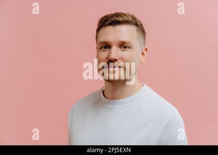 Serious handsome European man with stubble looks directly at camera dressed in casual white t shirt has calm face expression poses against pink backgr Stock Photo