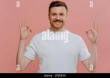 Young satisfied bearded man makes okay gesture with both hands expresses agreement approves something praises nice work says well done dressed in casu Stock Photo