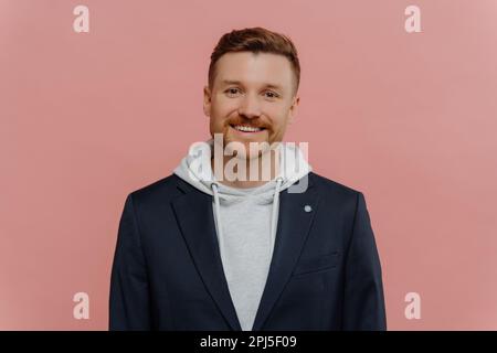 Indoor shot of handsome Caucasian man smiles toothily dressed in hoodie and black jacket has cheerful calm face expression enjoys pleasant talk poses Stock Photo