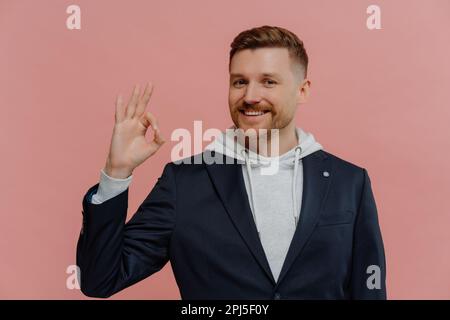 I agree. Young charming happy redhead man in casual wear showing ok gesture with fingers and smiling cheerfully at camera while posing isolated over p Stock Photo