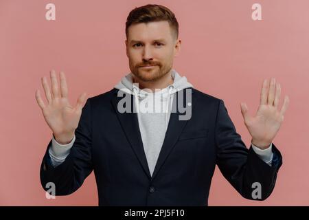 Serious confident adult man with bristle raises hands towards camera shows palms has strict expression asks to hold on or stop wears comfortable hoodi Stock Photo